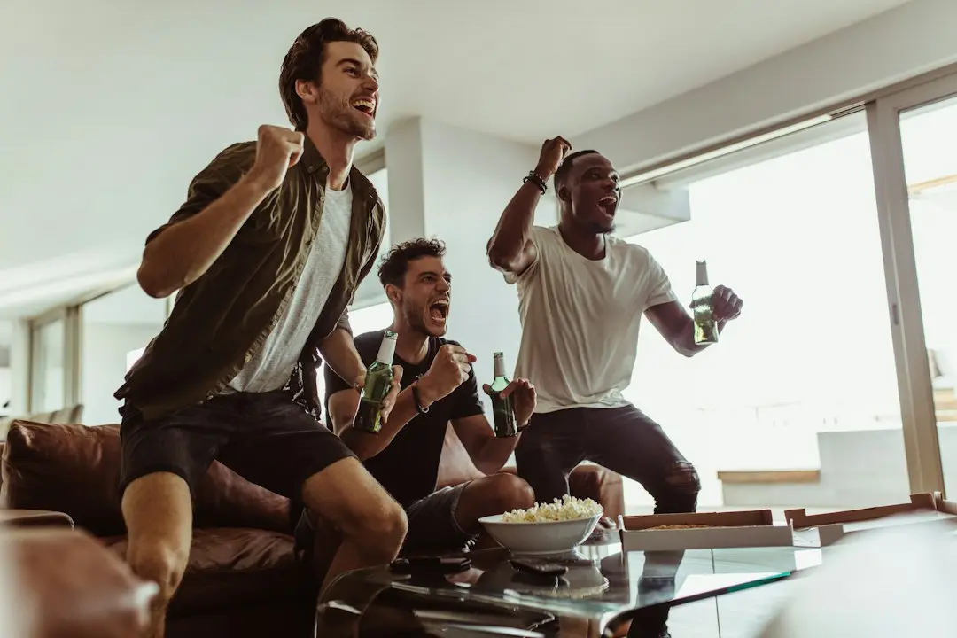 Tres amigos celebran de pie frente al sofá mientras ven un partido en casa
